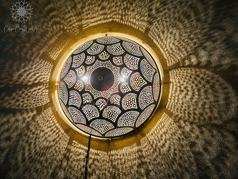 May include: A round, black and gold metal wall lamp with a detailed, patterned design. The lamp casts intricate light patterns on the surrounding wall. The lamp is mounted on a wall and has a power cord.
