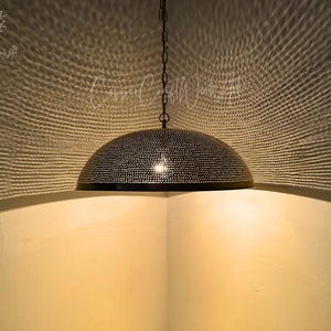 May include: A black, dome-shaped pendant light with a perforated design hangs from a chain. The light casts intricate patterns on the surrounding walls. The fixture is suspended in a corner, creating a warm, inviting ambiance. The overall theme is home decor.