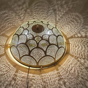 May include: A decorative, dome-shaped light fixture with a patterned design of small, evenly spaced holes. The light casts intricate shadows on the surrounding surface, creating a warm, inviting ambiance. The fixture is a warm gold color.