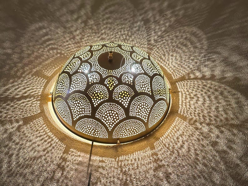 May include: A decorative, dome-shaped light fixture with a patterned design of small, evenly spaced holes. The light casts intricate shadows on the surrounding surface, creating a warm, inviting ambiance. The fixture is a warm gold color.
