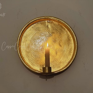 May include: A circular gold-colored metal wall sconce featuring a hammered texture on the reflective surface. A single cylindrical candle holder is attached to the bottom center, holding a lit off-white taper candle that casts a warm glow.