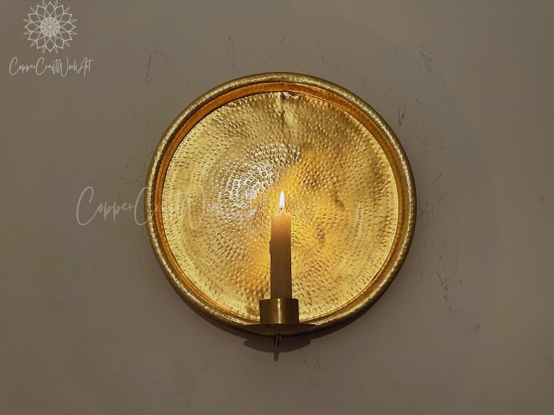 May include: A circular gold-colored metal wall sconce featuring a hammered texture on the reflective surface. A single cylindrical candle holder is attached to the bottom center, holding a lit off-white taper candle that casts a warm glow.