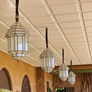 May include: Four hanging lanterns with white glass panels and dark metal frames suspended from a ceiling. Each lantern features a geometric, multi-faceted design with a pointed top and bottom, hanging by a dark strap from the ceiling structure.