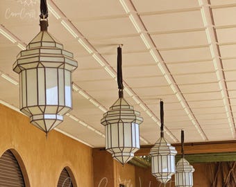 May include: Four hanging lanterns with white glass panels and dark metal frames suspended from a ceiling. Each lantern features a geometric, multi-faceted design with a pointed top and bottom, hanging by a dark strap from the ceiling structure.