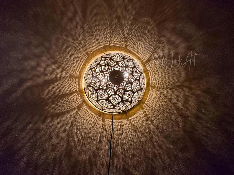 May include: A round, gold-colored, Moroccan-style ceiling light fixture with intricate cut-out patterns. The light casts a decorative pattern on the surrounding dark wall. A black cord extends from the bottom of the fixture.