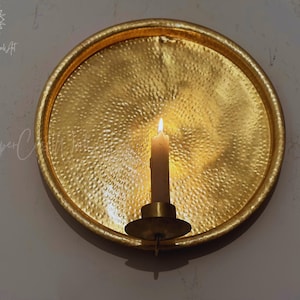 May include: A circular, gold-colored metal wall sconce featuring a hammered, textured surface. A single cylindrical candle holder is attached to the center, holding a lit, off-white taper candle that casts a warm glow across the metallic plate.