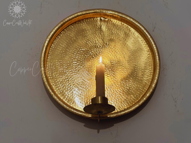 May include: A circular, gold-colored metal wall sconce featuring a hammered, textured surface. A single cylindrical candle holder is attached to the center, holding a lit, off-white taper candle that casts a warm glow across the metallic plate.