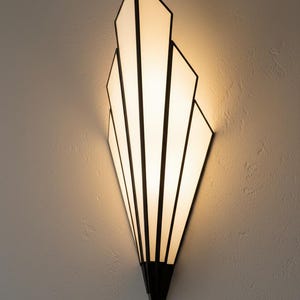 May include: A wall sconce featuring a fan-shaped design with five vertical panels of frosted white glass. The panels are framed by thin, dark metal lines that converge at a pointed base, creating a geometric silhouette that emits a warm glow.