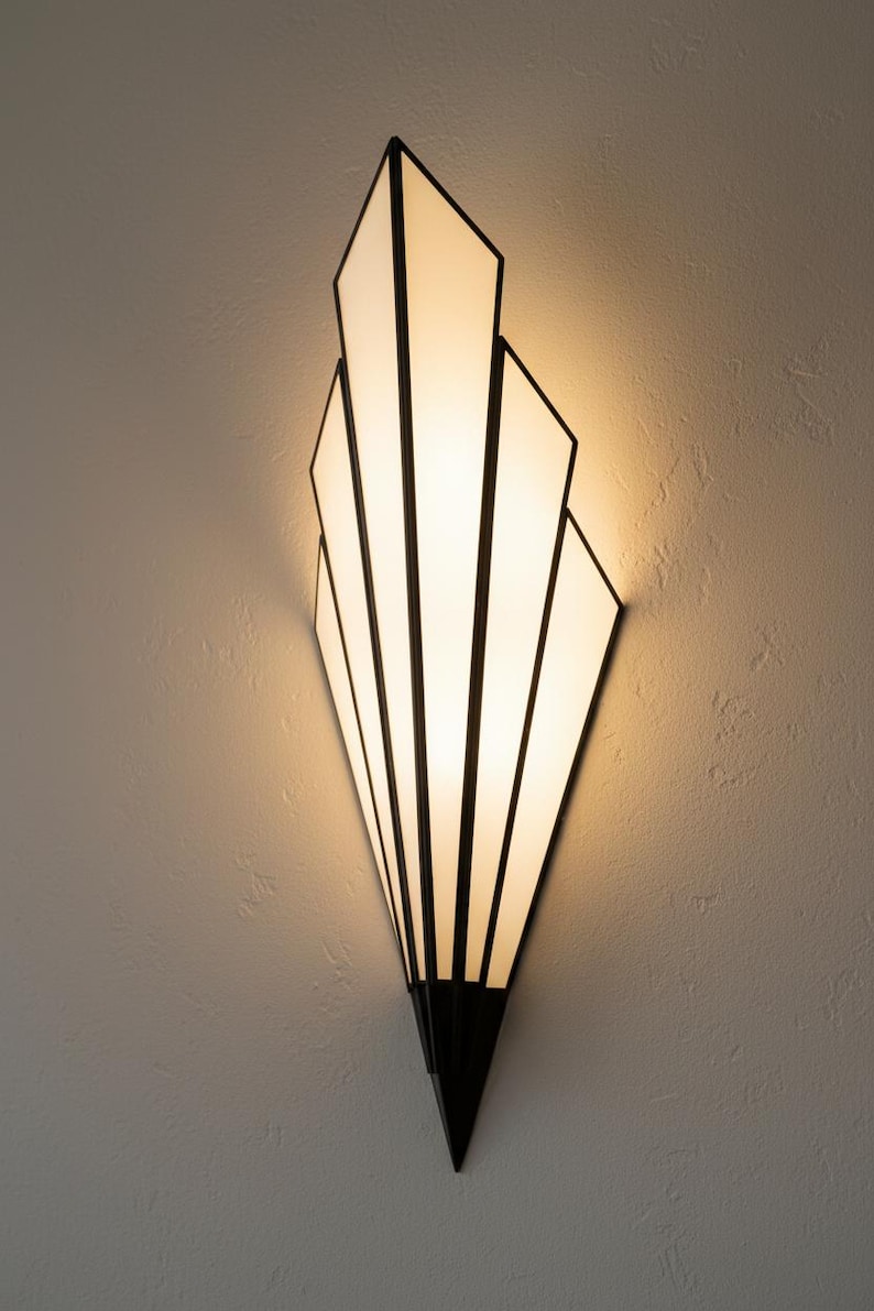 May include: A wall sconce featuring a fan-shaped design with five vertical panels of frosted white glass. The panels are framed by thin, dark metal lines that converge at a pointed base, creating a geometric silhouette that emits a warm glow.