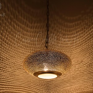May include: A round, metallic pendant light fixture with a dark finish and a perforated surface pattern. The lamp is suspended by a dark chain and emits a warm, glowing light through its numerous small holes, casting a textured shadow pattern.