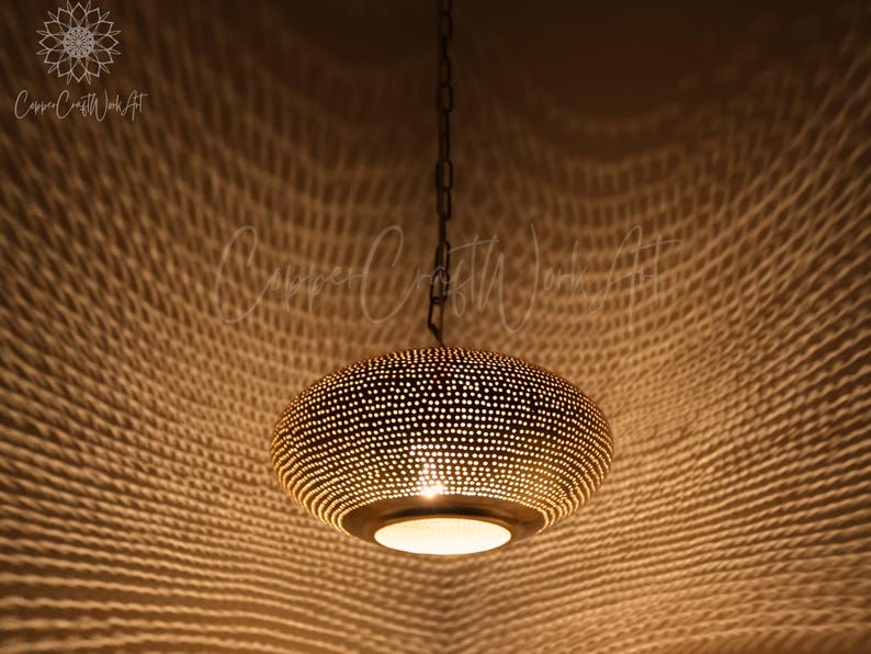 May include: A round, metallic pendant light fixture with a dark finish and a perforated surface pattern. The lamp is suspended by a dark chain and emits a warm, glowing light through its numerous small holes, casting a textured shadow pattern.
