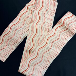 May include: Cream-colored fabric with a repeating wavy pattern of red, pink, and gray stripes. The fabric is folded and arranged on a dark blue surface. The pattern creates a flowing, dynamic visual effect.