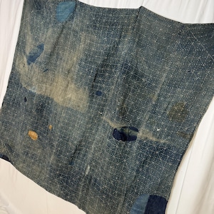 May include: A rectangular indigo-dyed textile featuring a dense, white geometric sashiko-style stitched grid pattern. The fabric displays visible signs of wear, including multiple patches in varying shades of blue and tan, along with frayed areas.