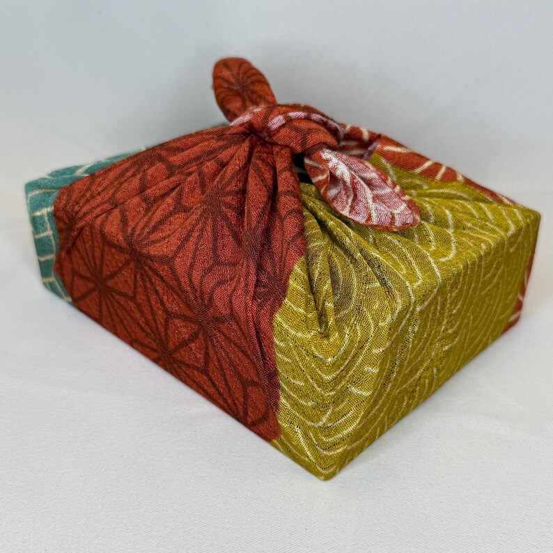May include: A gift box wrapped in patterned fabric. The fabric is in shades of red, green, and yellow, with a geometric design. The fabric is tied at the top with a knot, creating a decorative and eco-friendly presentation.