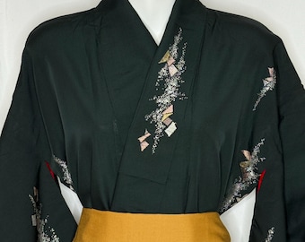 Op de afbeelding: Een zwarte kimono met een mosterdkleurige sjerp. De kimono heeft geborduurde bloemen- en geometrische ontwerpen in roze, wit en goud. De mouwen zijn lang en het kledingstuk wordt op een mannequin getoond.