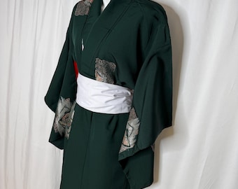 Op de afbeelding: Een donkergroene kimono met een witte obi-riem. De kimono heeft patronen op de mouwen en kraag. Het kledingstuk wordt op een mannequin gepresenteerd.