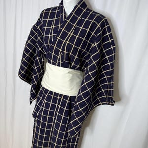 Op de afbeelding: Marineblauwe kimono met een crèmekleurig rasterpatroon. De kimono heeft wijde mouwen en wordt in de taille vastgehouden met een brede, crèmekleurige obi-riem. Het kledingstuk wordt op een mannequin gepresenteerd.