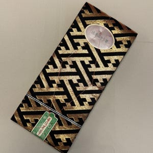 May include: A rectangular package with a black and gold geometric pattern. The package has a tag with Japanese text and a small oval sticker with Japanese text. The package is tied with a copper-colored string.