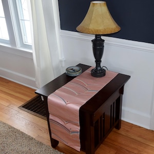 May include: A dark wood end table with a pink and white patterned table runner. A lamp with a brown lampshade sits on the table.