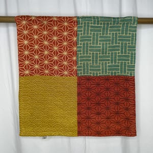 May include: A patterned textile with a geometric design. The fabric is divided into four sections, each with a different pattern in red, green, and gold. The designs include starbursts, woven lines, and wave-like shapes.