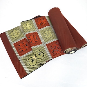 May include: A rolled-up textile with a repeating floral pattern in squares. The squares alternate between yellow and orange flowers on a white grid. The border and rolled section are a solid dark red.
