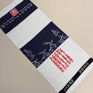 May include: A rectangular white and navy blue Japanese towel with a fringed edge. The towel features Japanese characters and illustrations of waves and figures. The top and bottom edges have black bands with Japanese text.