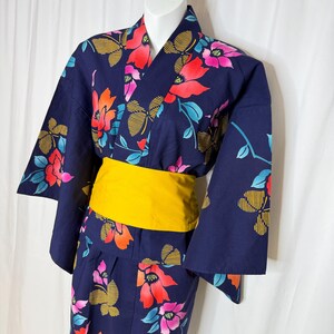 May include: A dark blue yukata featuring a vibrant floral pattern with red, pink, and orange blossoms, accented by yellow striped butterfly motifs. The garment is cinched at the waist with a solid, bright yellow obi sash, showcasing wide sleeves.