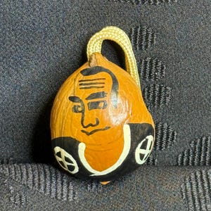 May include: A hand-painted, gourd-shaped ornament featuring a stylized face with black and white accents. The ornament has a yellow cord for hanging. The face is painted on an orange background with black and white details.