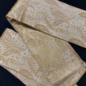 Puede incluir: Primer plano de una tela color crema con un estampado de cachemira dorado. La tela está doblada, revelando el intrincado diseño y la textura. El estampado de cachemira está detallado con hilos metálicos dorados, creando un aspecto lujoso.