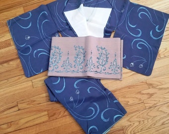 Op de afbeelding: Een blauwe en witte kimono met een roze en witte band. De kimono heeft een traditioneel Japans ontwerp met een bloemenmotief.