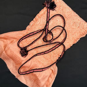 May include: A peach-colored scarf with a geometric pattern is paired with a black and pink rope belt. The belt has pink stitching and black edges, with tassels at each end. The scarf is folded and draped on a black surface.