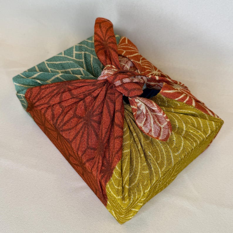 May include: A square package wrapped in a colourful cloth using the Furoshiki method. The fabric features a patchwork design with teal, red, and yellow-green sections, each with a different pattern. The knot is at the top, securing the package.