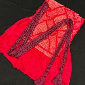 May include: A vibrant red textile with a geometric pattern is paired with two long, braided burgundy cords, each ending in a tassel. The cords are intricately woven, adding texture to the design. The overall aesthetic is bold and eye-catching.