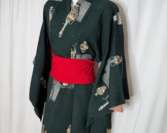 Op de afbeelding: Donkergroene kimono met een rode obi-riem. De kimono heeft een herhalend patroon van gestileerde vazen en andere decoratieve elementen in tinten beige, grijs en geel. De mouwen zijn wijd en het kledingstuk is aan de voorkant open.