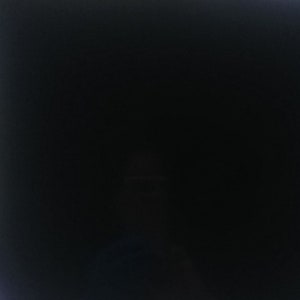 May include: A large, square, black object is displayed on a decorative metal stand. The object is reflective, and the background is blurred, showing a window and outdoor foliage. The stand has a simple, geometric design.