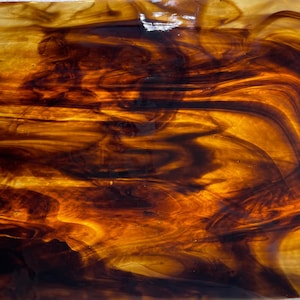 May include: A rectangular piece of stained glass with a swirling pattern of amber and dark brown hues. The glass has a glossy finish and is set against a textured, silver-colored background. The design evokes a sense of warmth and depth.