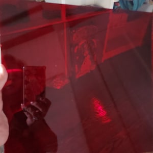 May include: A transparent, rectangular sheet of deep red plastic. The sheet is held up, reflecting light and the surrounding environment. The edges of the sheet are visible, and the overall effect is a vibrant, glossy appearance.