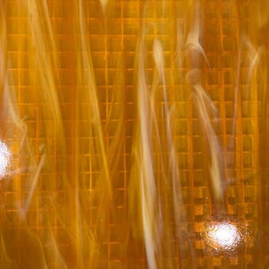 May include: Close-up of a textured, amber-colored glass sheet. The glass has a grid pattern overlaid with flowing, white and amber swirls, creating a dynamic and artistic effect. The surface is glossy and reflects light.