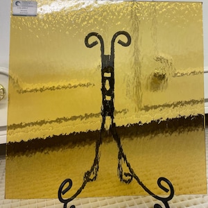 May include: A square piece of textured glass with a gold color. The glass is on a black stand.