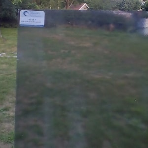May include: A large, rectangular sheet of pale gray glass, labeled "180-85-F Pale Gray Solid Transparent." The glass is partially transparent, with a slight tint of gray. The background shows green grass and trees.