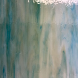 May include: A sheet of stained glass with a flowing pattern of teal, white, and light green. The colors blend together, creating a marbled effect. The glass has a glossy finish and appears to be translucent, allowing light to pass through.