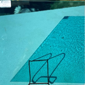 May include: A square, transparent, teal-colored glass panel with a black square border and a shadow of a square shape. The panel is placed over a light teal surface, with a darker teal surface in the background.