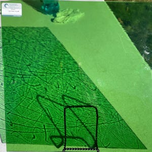 May include: A collection of green glass sheets, with varying shades and textures. One sheet has a textured pattern of lines, while others are smooth. A black metal stand is in the foreground. The image includes a label with text.