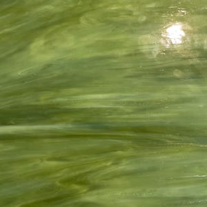 May include: A close-up of a sheet of green stained glass. The glass has a marbled appearance with varying shades of green and white, creating a flowing, organic pattern. The surface reflects light, highlighting the texture.