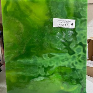 May include: A green glass sheet with a swirled pattern. The sheet is on a wooden stand. A label on the sheet reads "Youghighemy Glass (724) 828-3000 4504 SP Made in the USA"