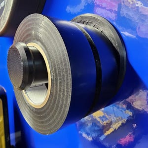 May include: Close-up of a roll of black electrical tape with a textured surface. The tape is mounted on a blue surface. The tape roll has a black plastic end cap.