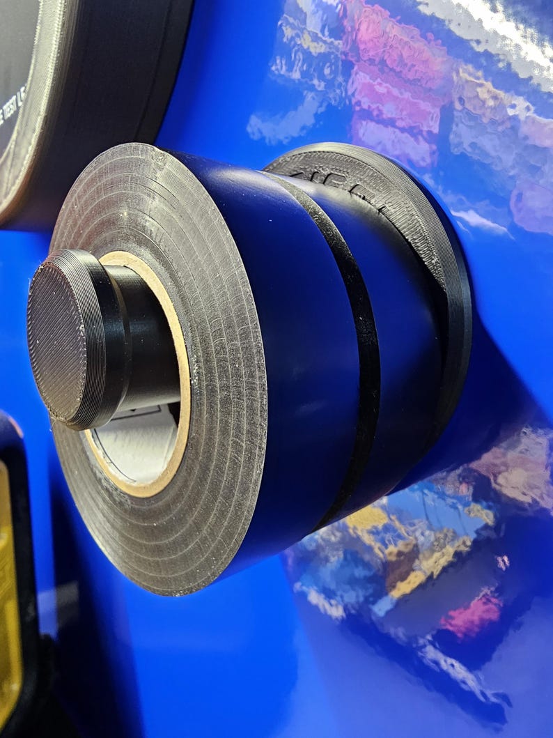 May include: Close-up of a roll of black electrical tape with a textured surface. The tape is mounted on a blue surface. The tape roll has a black plastic end cap.