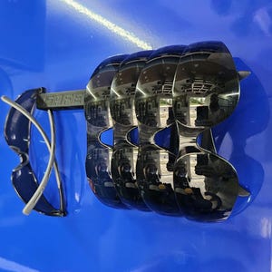 May include: A display of black sunglasses with a reflective surface, mounted on a blue background. The sunglasses are arranged in a row, with the word "SARE" visible on the mounting bar. One pair of sunglasses is positioned separately.
