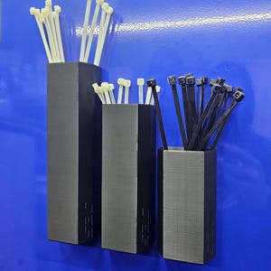 May include: Three matte black, hexagonal, wall-mounted storage containers of varying heights. The two taller containers hold white plastic cable ties, while the shortest container holds black plastic cable ties. Each container features the text "DES" embossed.