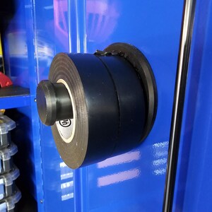 May include: A roll of black electrical tape mounted on a blue surface. The tape is held in place by a black holder. The tape roll has a white label with a logo. A metal rod is visible in the background.